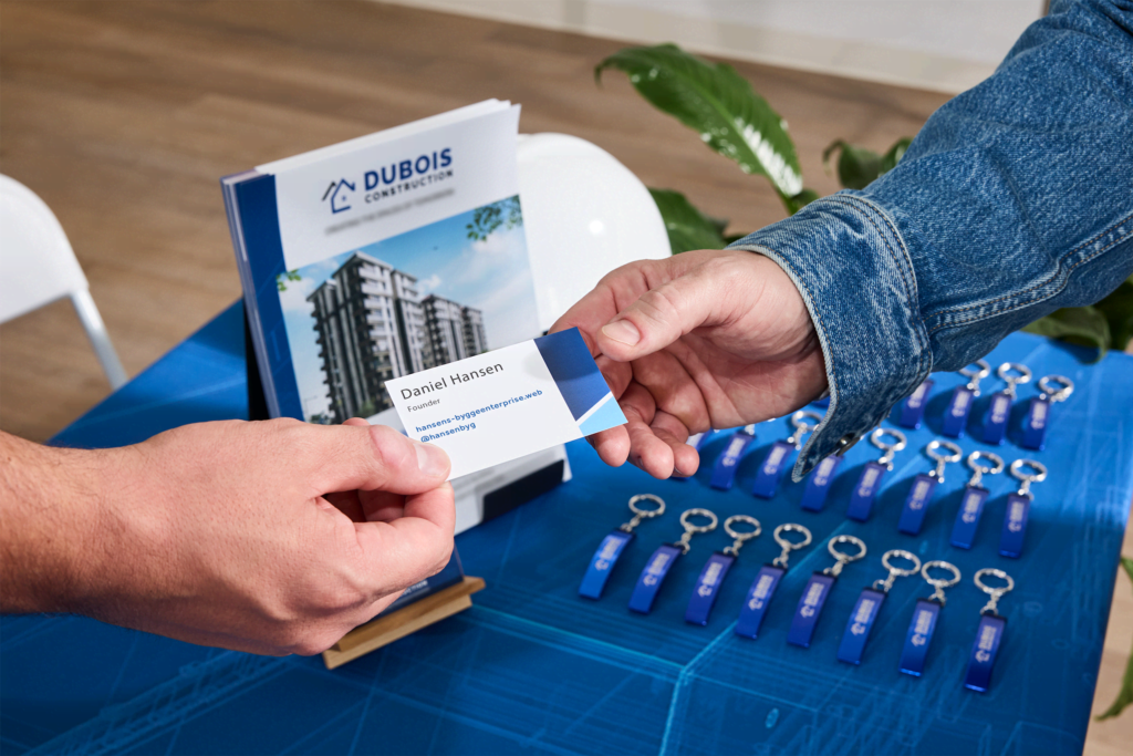 Small business owner handing a business card to another person at a trade show table with Dubois Construction brochures and blue promotional keychains.
