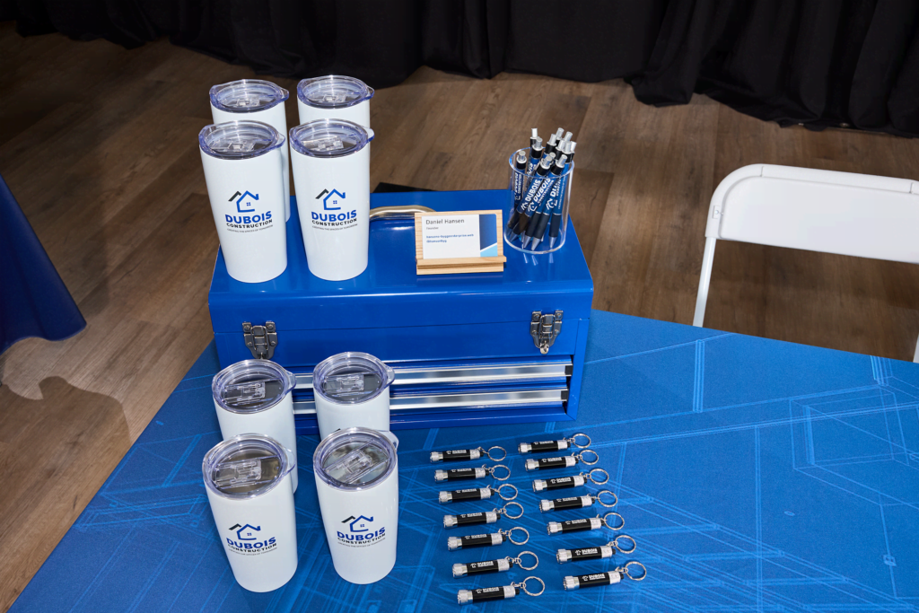 A trade show display table with Dubois Construction branded tumblers, business cards, roller banners, and promotional keychains on blue tablecloth.
